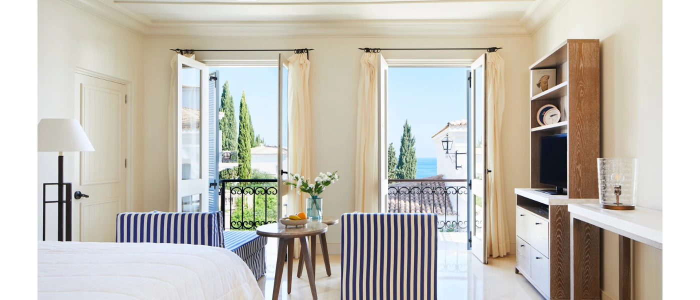 Stylish hotel room with blue and white pinstriped armchairs, a wooden bookcase with a TV, and a small wooden table overlooking a balcony with black wrought-iron railings and a a view of cypress trees and the sea