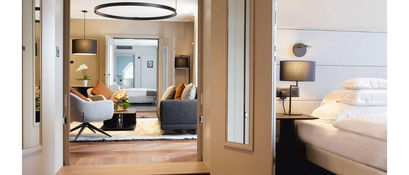 Corner of a white bed in a hotel room, with a doorway showcasing a living area with a sofa and chair in stylish grey and terracotta tones