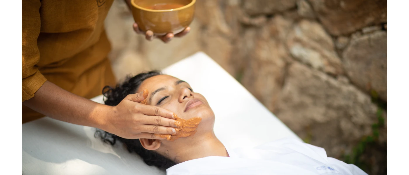 Woman lies on a bed in a white robe with her eyes closed as a therapist spreads an orange mask on her face
