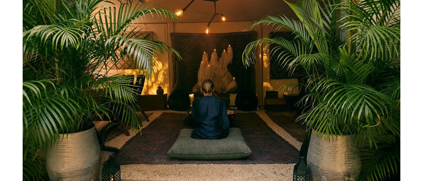 Female in a silk robe sits on an oversized floor cushion in a softly lit room, with two potted palms behind her