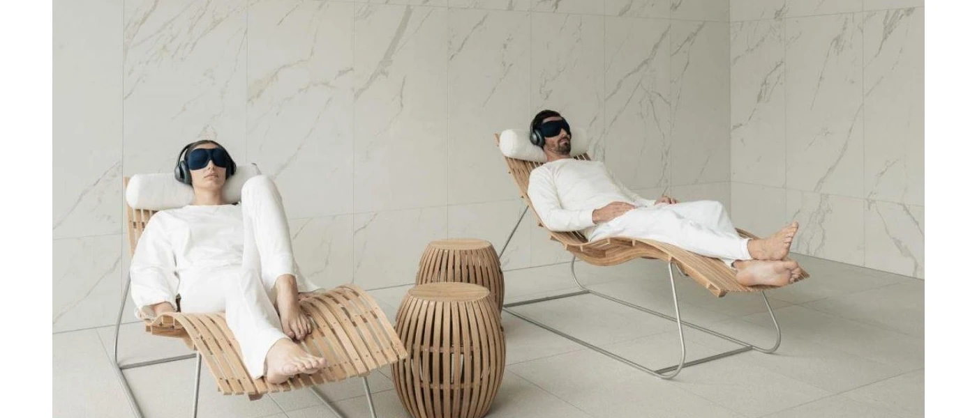 A man and woman in white clothes lying on wooden loungers wearing black eye masks and headphones, in a room with white marble walls