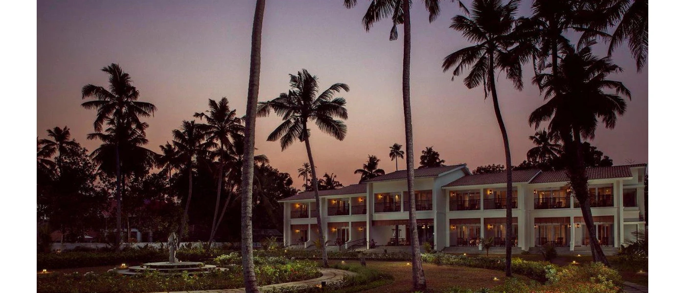 Two-storey white hotel with black balconies and surrounding palm gardens
