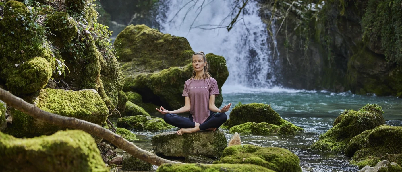 Woman in dark leggings and a purple T shirt meditating on a rock surrounded by other rocks and a waterfall