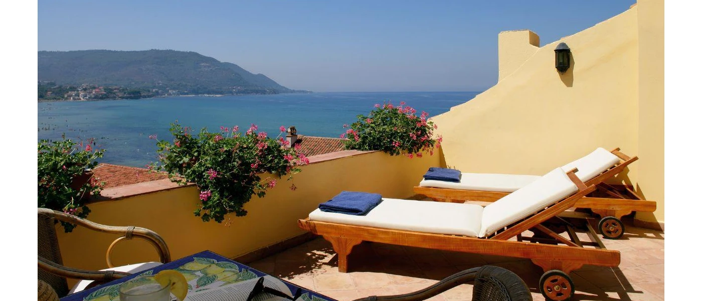 Sunny terrace with wooden white-cushioned loungers and blue towels, overlooking the sea and mountains 