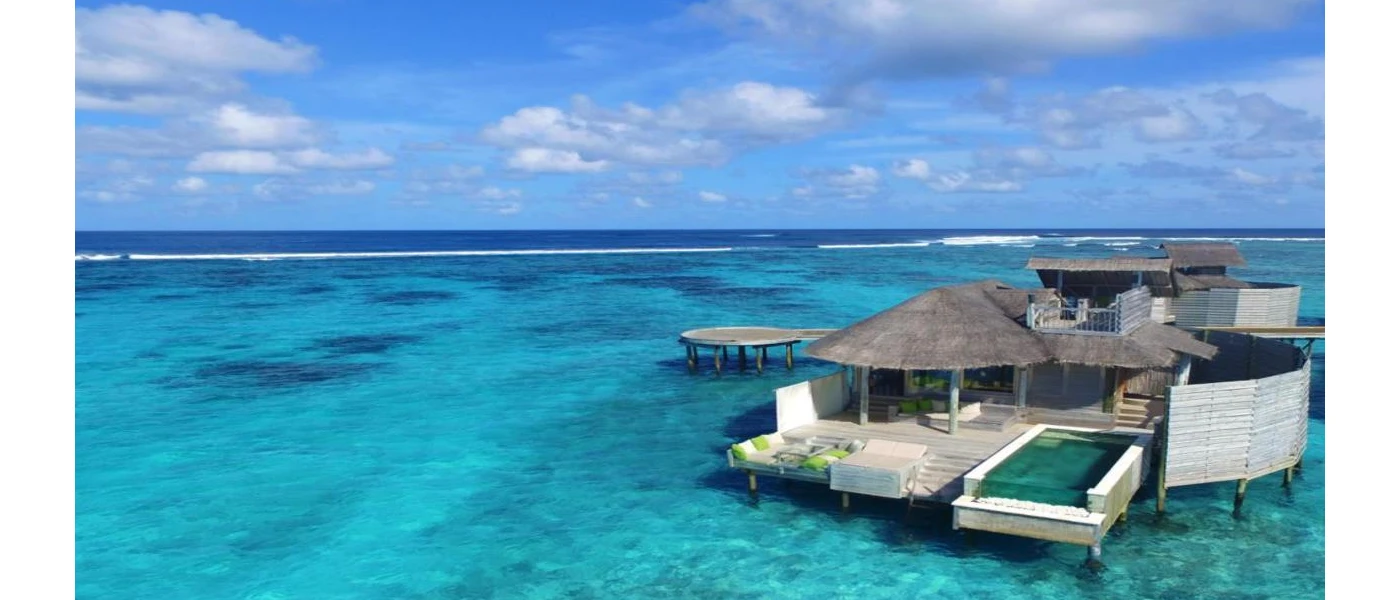 Thatched overwater villa in turquoise shallows, with a wooden deck with green cushions and a swimming pool