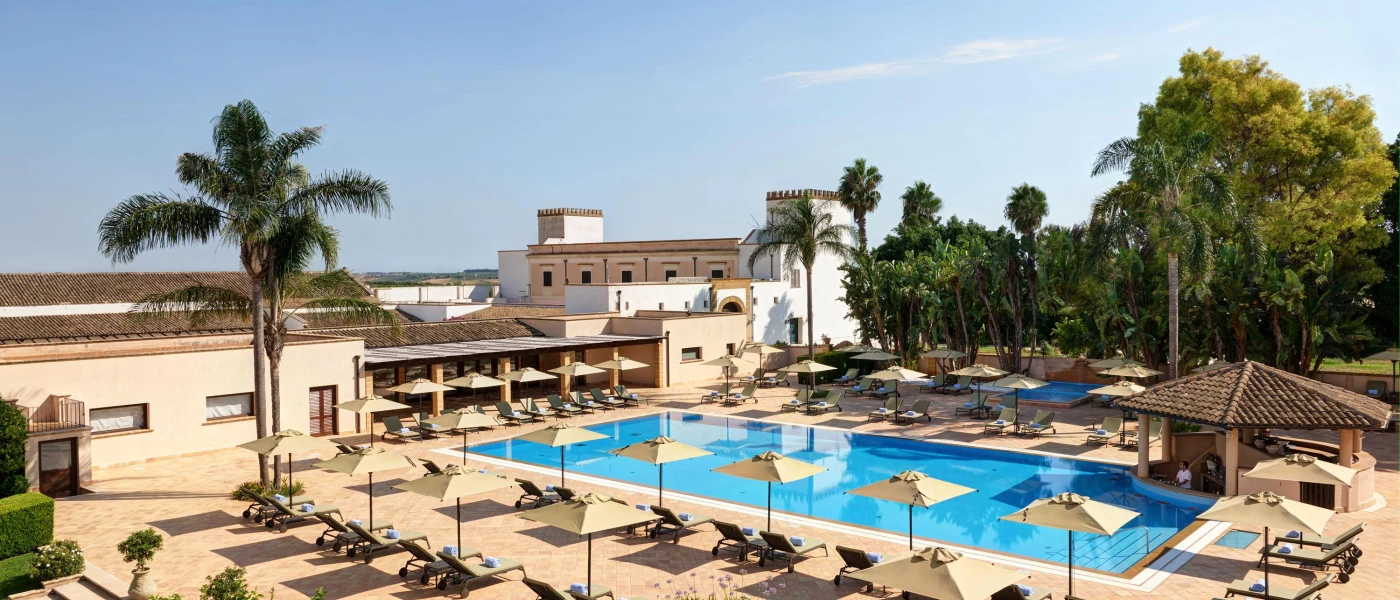 Rectangular pool in a stone terrace furnished with cream loungers and umbrellas, with a terracotta-roofed swim-up bar and surrounding trees