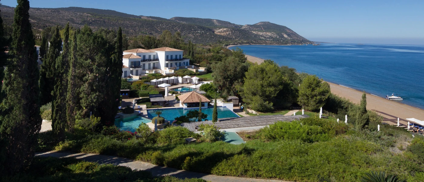 Cypress trees and lush shrubs cloaking a whitewashed resort and central swimming pool, with a gold sandy beach and blue sea in the background