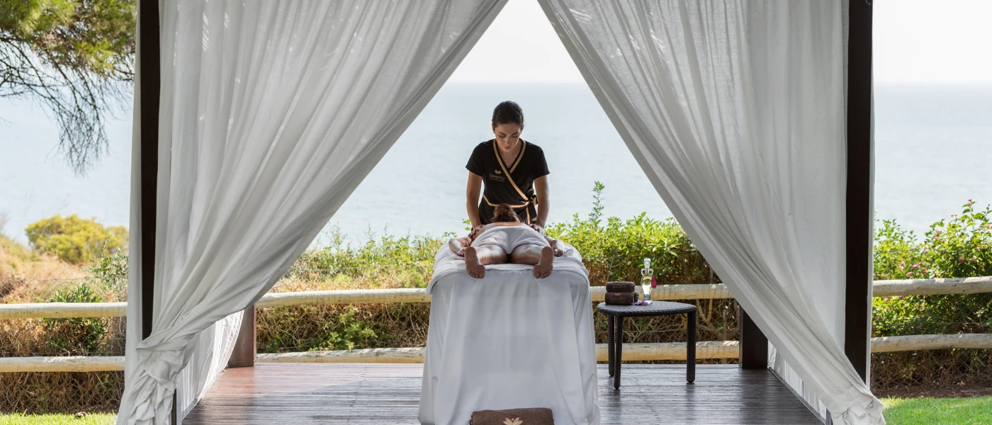 Woman lies on her front in an open-air white cabana in front of the ocean, while enjoying an outdoor massage