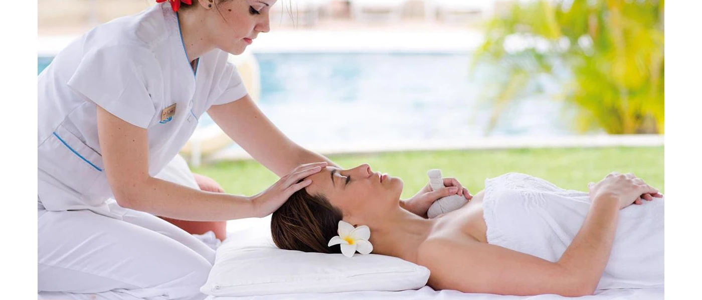Woman lies on her back in a white towel with a white flower in her hair, and a female therapist places her hands on her hand and chest