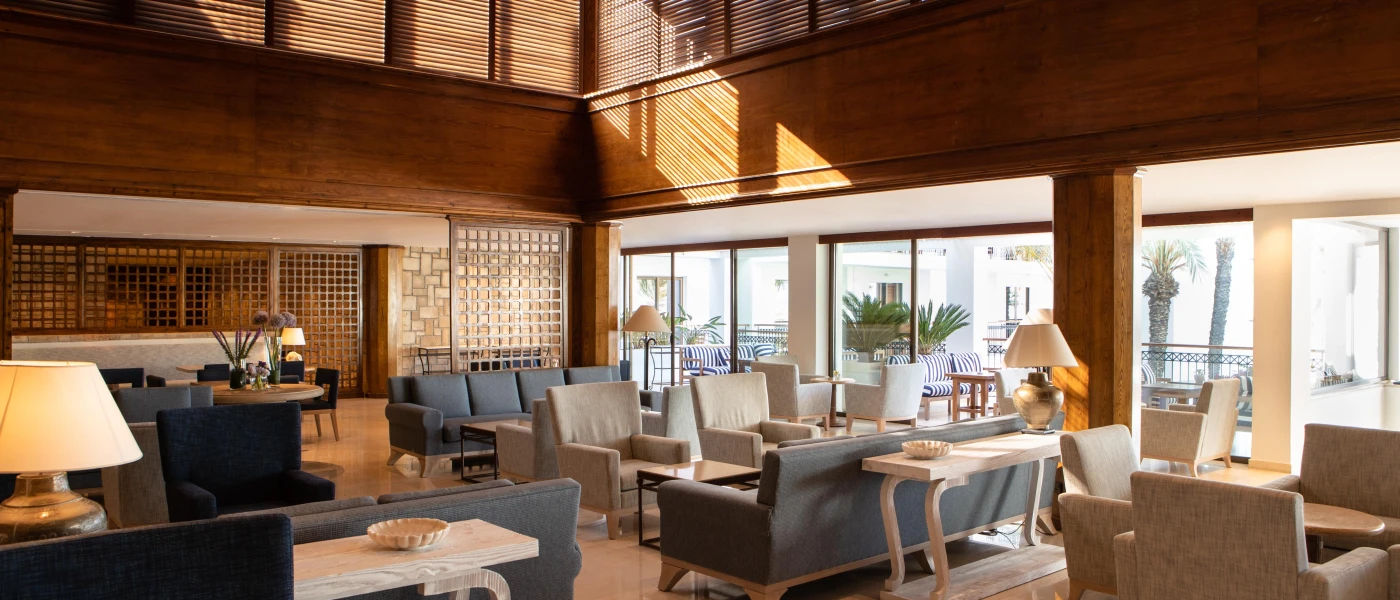 Airy lobby/lounge area, with wood-panelled walls, grey and stone chairs and sofas, and floor-to-ceiling windows overlooking a furnished terrace