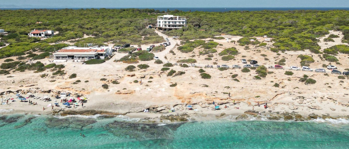 White hotel buildings overlooking a sandy beach and turquoise waves surrounded by pine forests