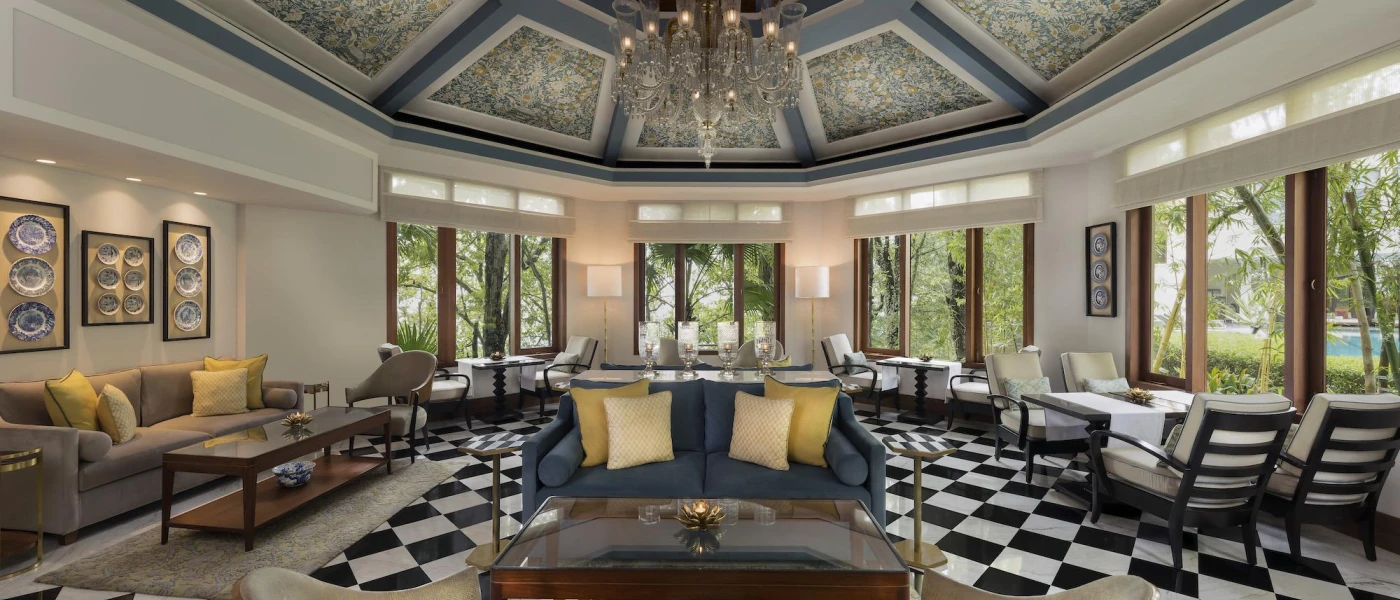 Lounge area with a black and white chequered floor, blue and stone-coloured sofas with yellow cushions, and windows with views of the greenery outside