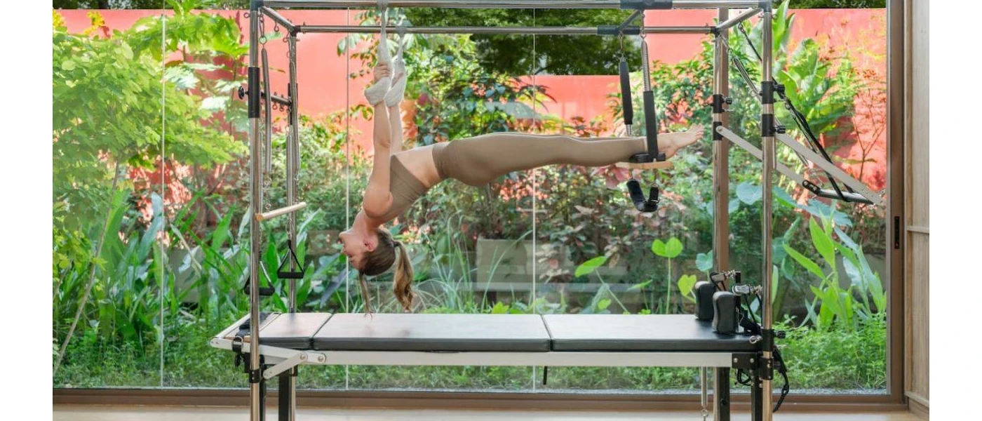 Woman in brown active wear stretches in a reformer Pilates machine in front of tropical plants