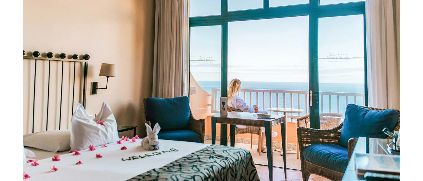 Hotel room with cream walls, white bedding with pink rose petals, a blue armchair and a balcony where a woman sits looking out to sea