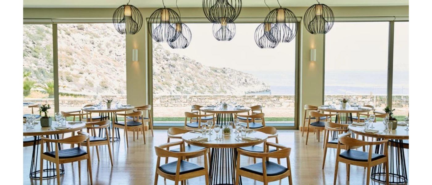 Airy restaurant with floor-to-ceiling windows with views of the sea and craggy cliffs, simple wooden tables and chairs and a cluster of contemporary ceiling pendant lights in a wiry black design