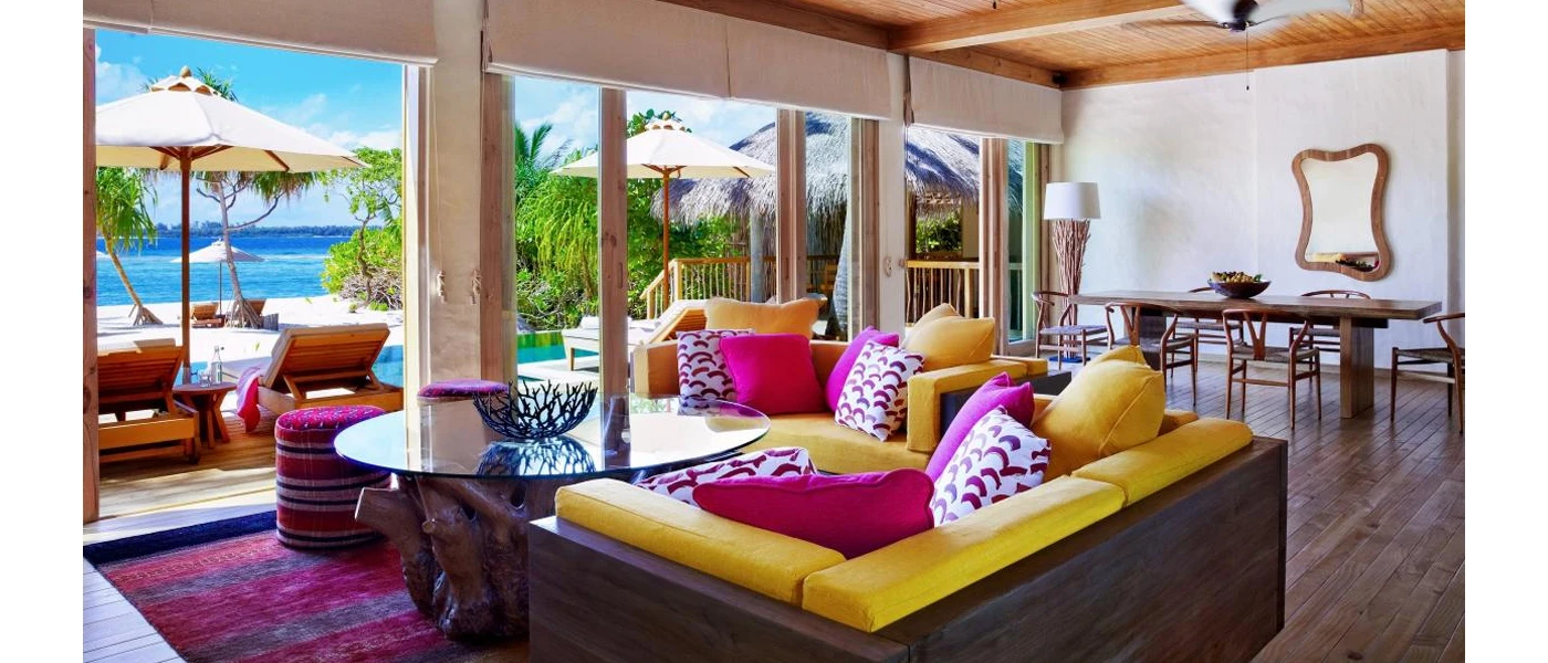 Villa interior with a dark-wood sofa with yellow and pink cushions, a glass coffee table with a driftwood-design bottom, a dining table behind, wood-beamed ceiling and full-length windows overlooking the beach and ocean