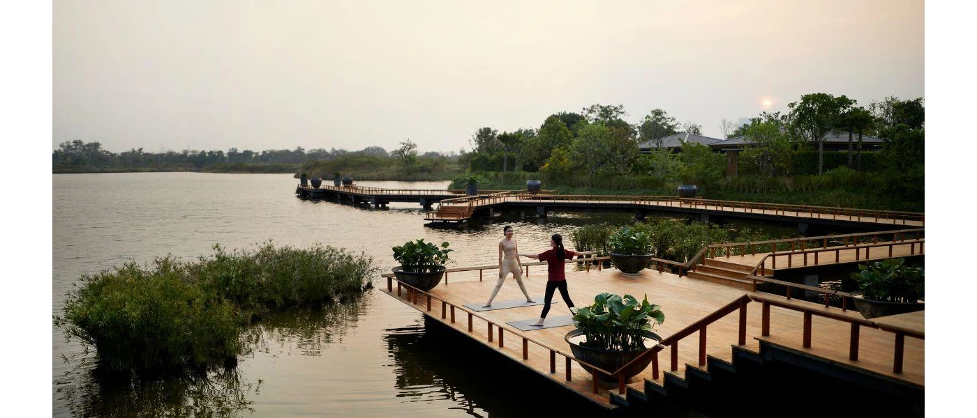 Two women in active wear stand on a wooden deck on a riverfront surrounded by greenery