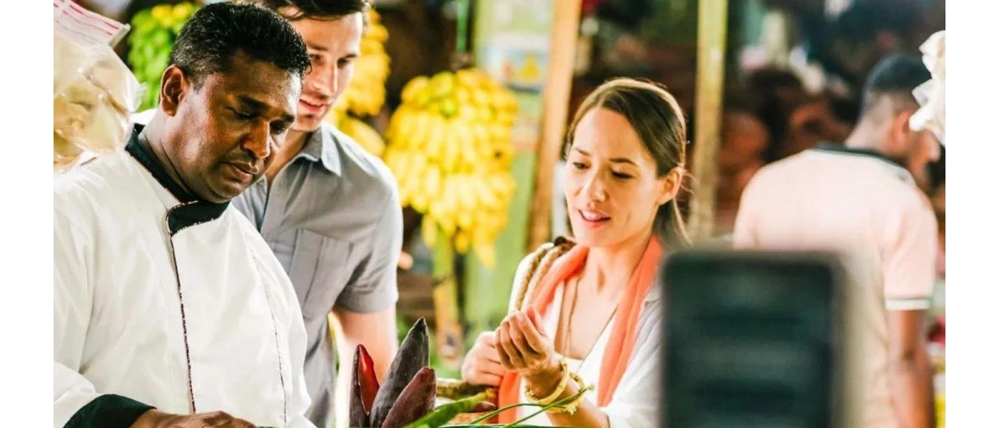 Chef in whites preparing ingredients as two guests watch on smiling