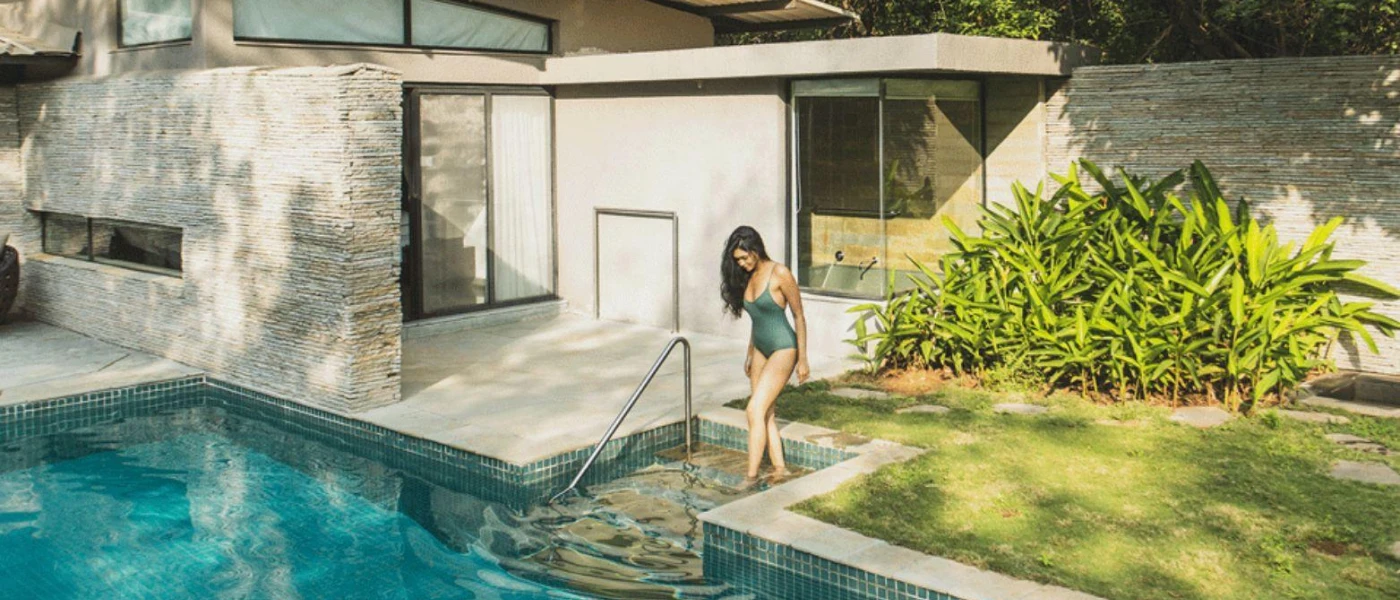 Woman with long dark hair in a dark swimsuit steps into a pool outside a contemporary villa