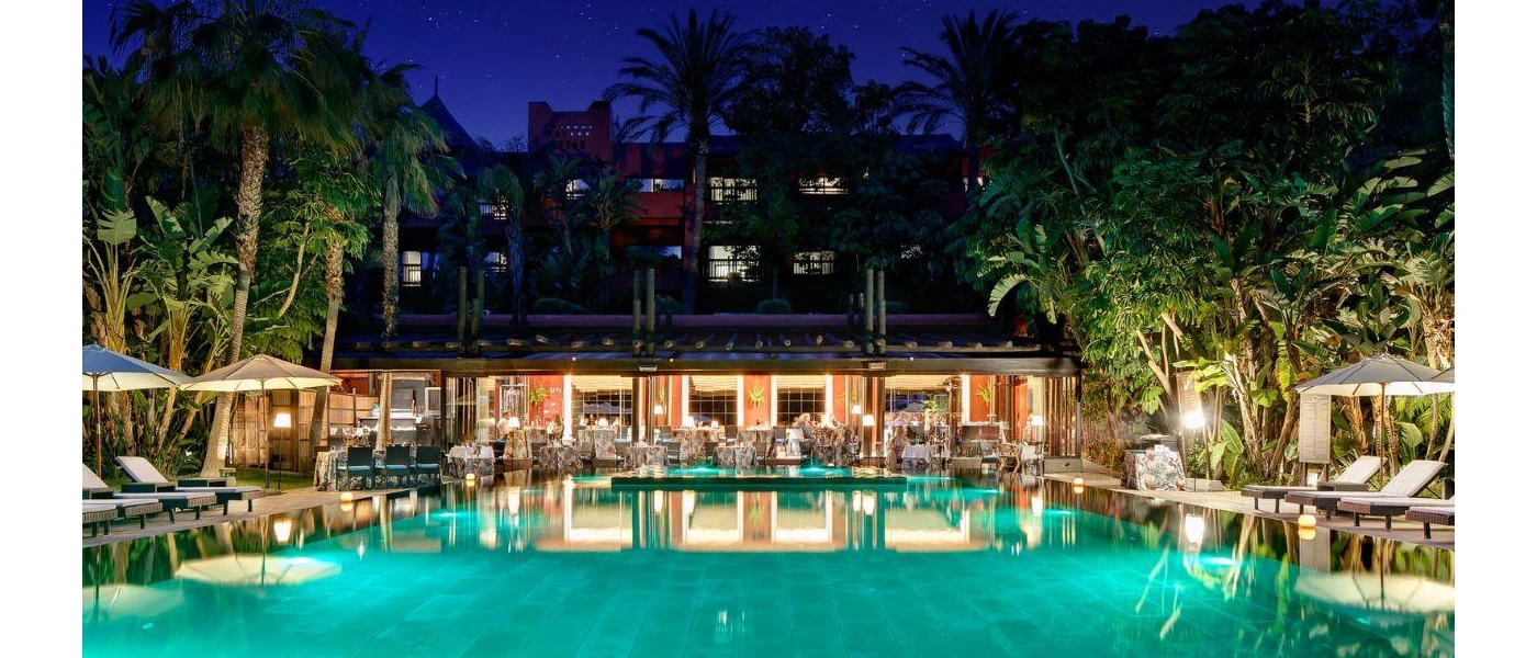 Night-time at an outdoor pool, surrounded by white loungers and umbrellas and tropical greenery, in front of a al fresco restaurant terrace