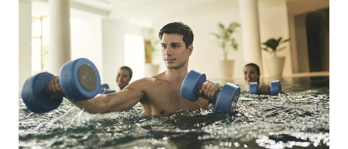 Man exercises with blue dumbbells in an indoor swimming pool with two people behind him