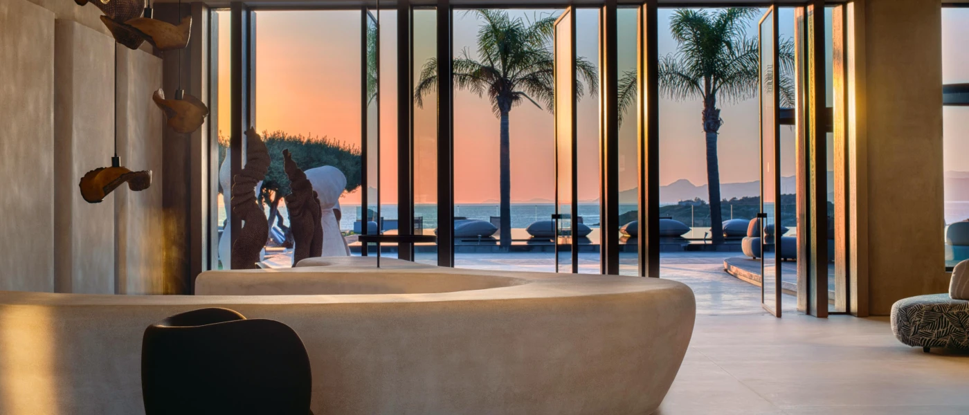 Reception area with cream limewashed walls, a large marble-like cream desk and a brass ceiling light cluster, with a view to a palm-lined swimming pool