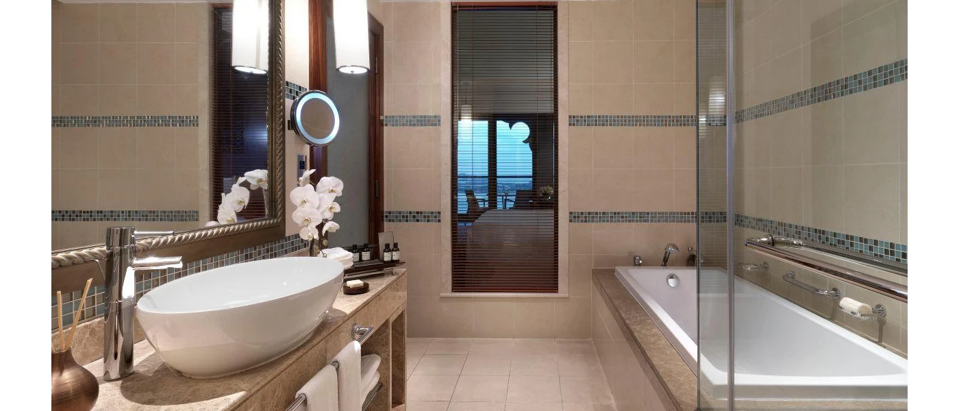 Neutral tiled bathroom with marble finishes, a rectangular bathtub, vanity unit with a large white sink, glass bottled amenities and a vase of orchids