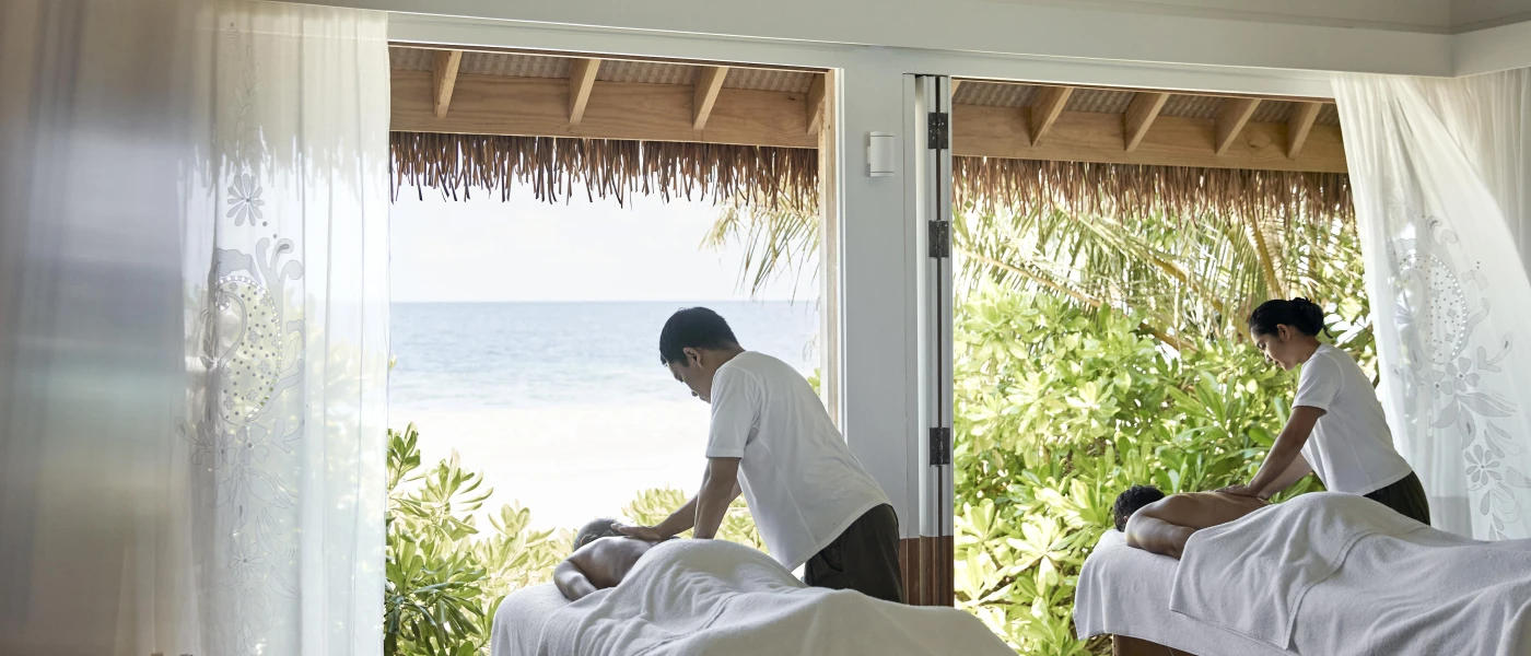 Two people enjoy a massage on neighbouring beds in an open-sided cabana with a view of the ocean