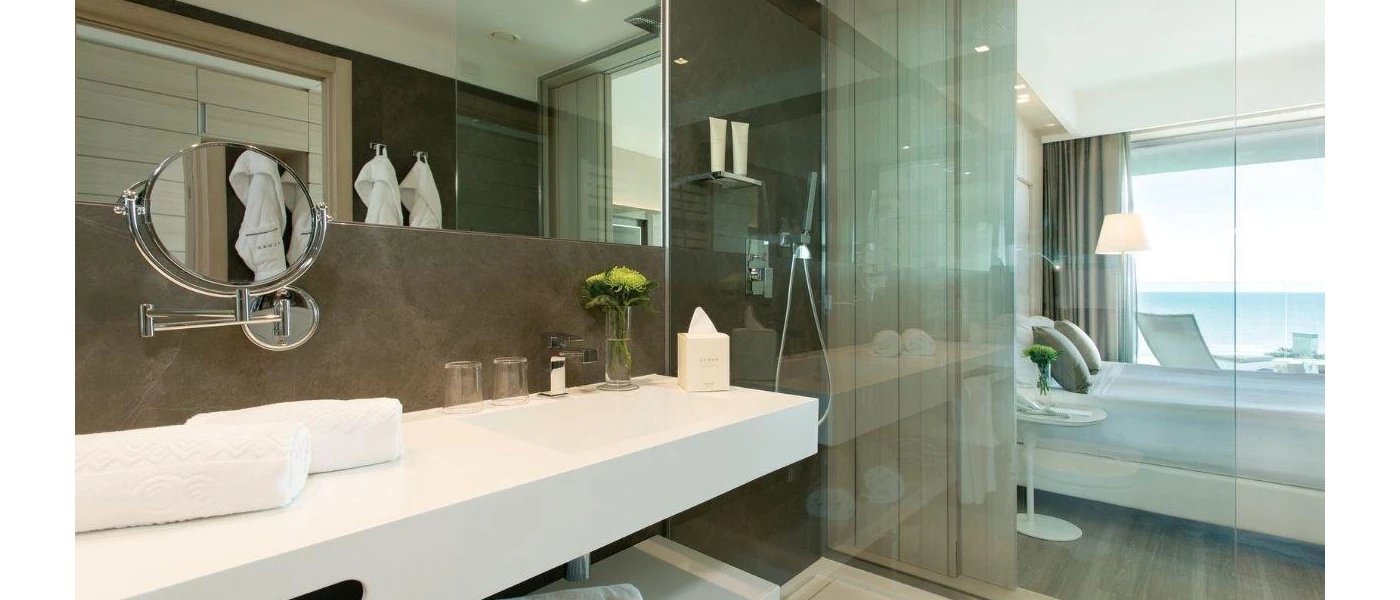 Sleek hotel bathroom with a marble wall, glass wall showing the bedroom, white long vanity unit and robes hanging up