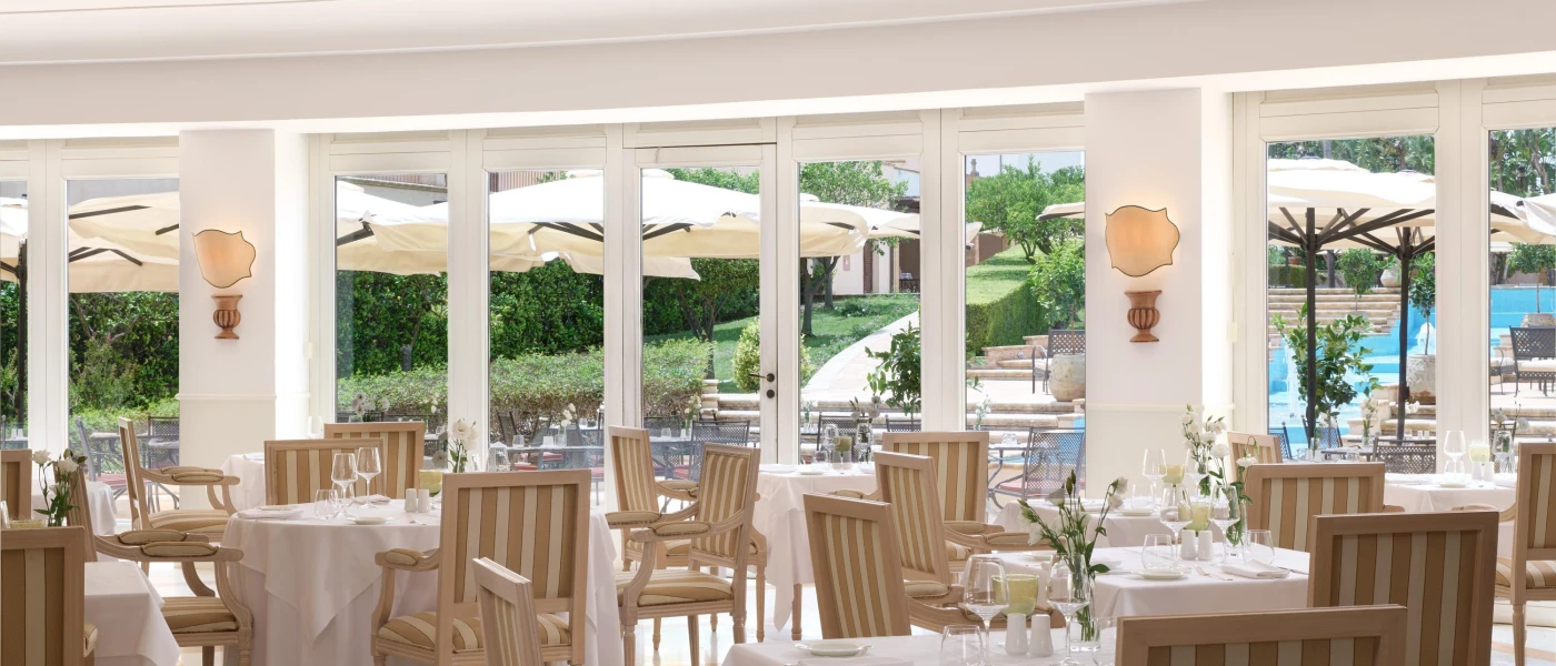 Airy restaurant with white tablecloths, wooden chairs, full-length windows and a view of an outdoor fountain and gardens
