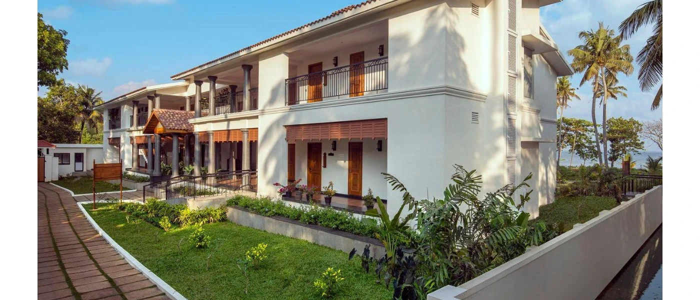 White two-storey building with grey pillars, black balconies and surrounding tropical gardens