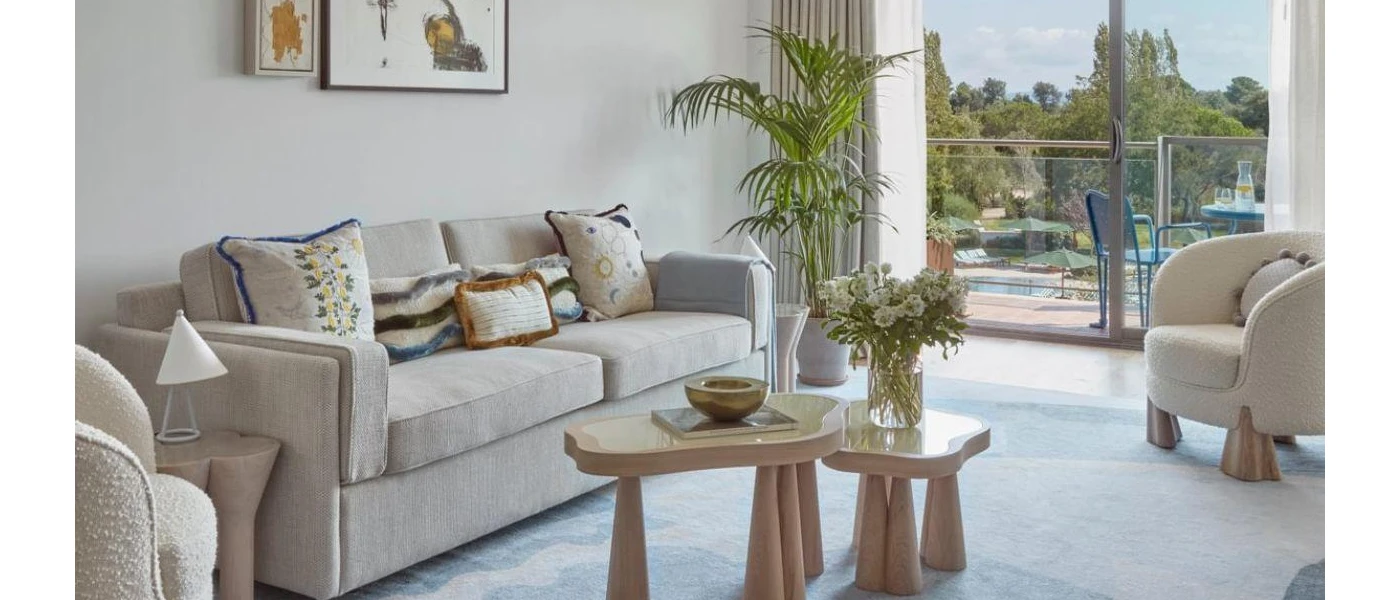 Hotel room living area with neutral sofas, two small wooden coffee tables, patterned cushions and a rug, plus floor-to-ceiling balcony doors