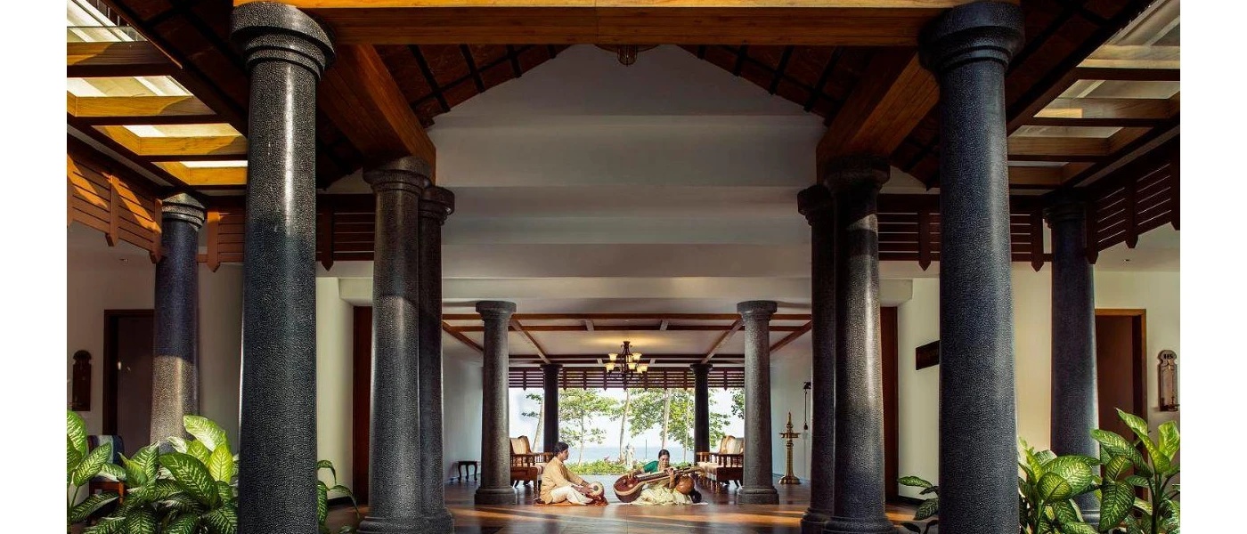 Airy lobby area with plants and dark grey pillars, with two Indian musicians sat playing instruments on the floor