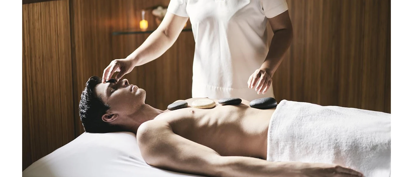 Man lies on a bed with a white towel covering his bottom half as a therapist lines stones down his body