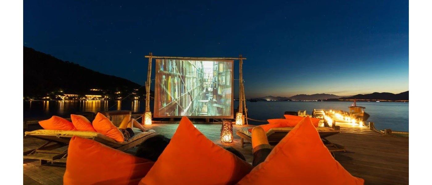 Large cinema screen in front of wooden loungers with orange cushions, next to a wooden jetty on the seafront under a night sky