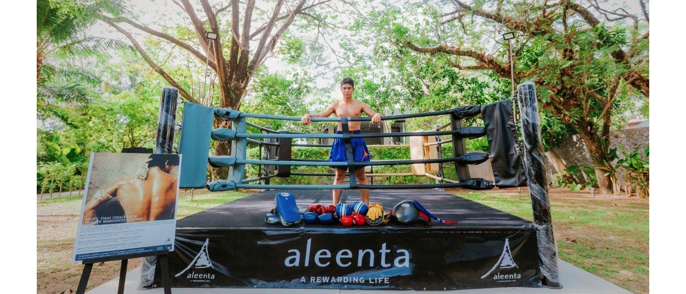 Muay Thai boxer stands in a ring in blue shorts in a leafy garden, with a sign next to him detailing 'private sessions on request'