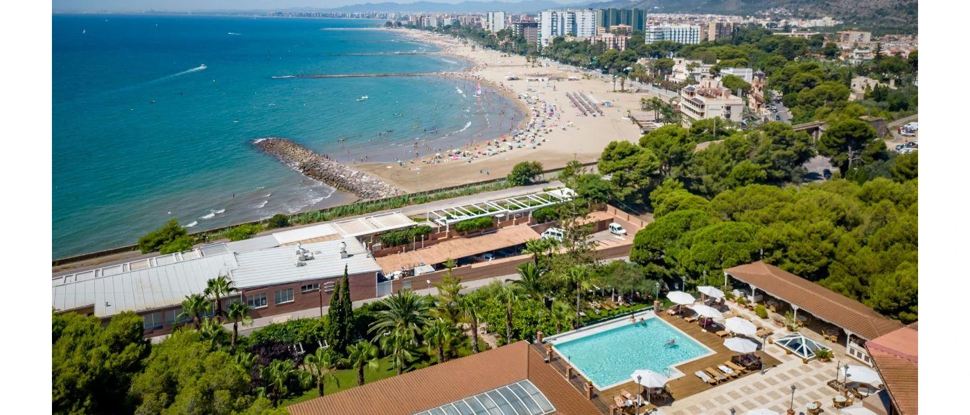 Sunny terrace with a pool, loungers and umbrellas surrounded by greenery overlooking a sandy beach, the sea and a high-rise town