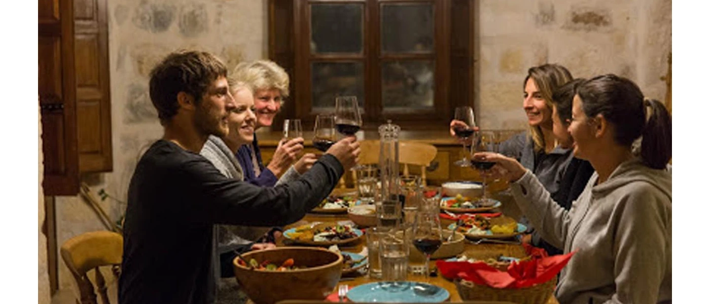 Smiling group clink glasses of red wine in a stone-walled room over a table filled with colourful food