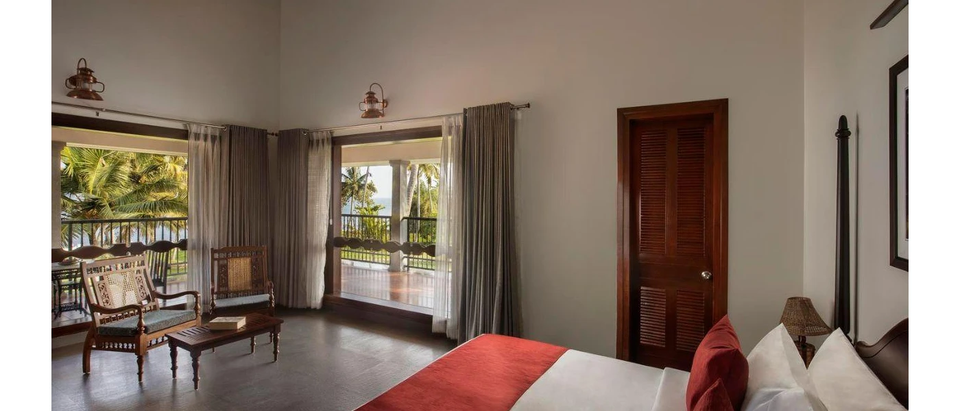 Hotel room with white walls, tiled flooring, white bedding with red cushions and a throw, and dark-wood furnishings, with floor-to-ceiling windows and doors overlooking a balcony