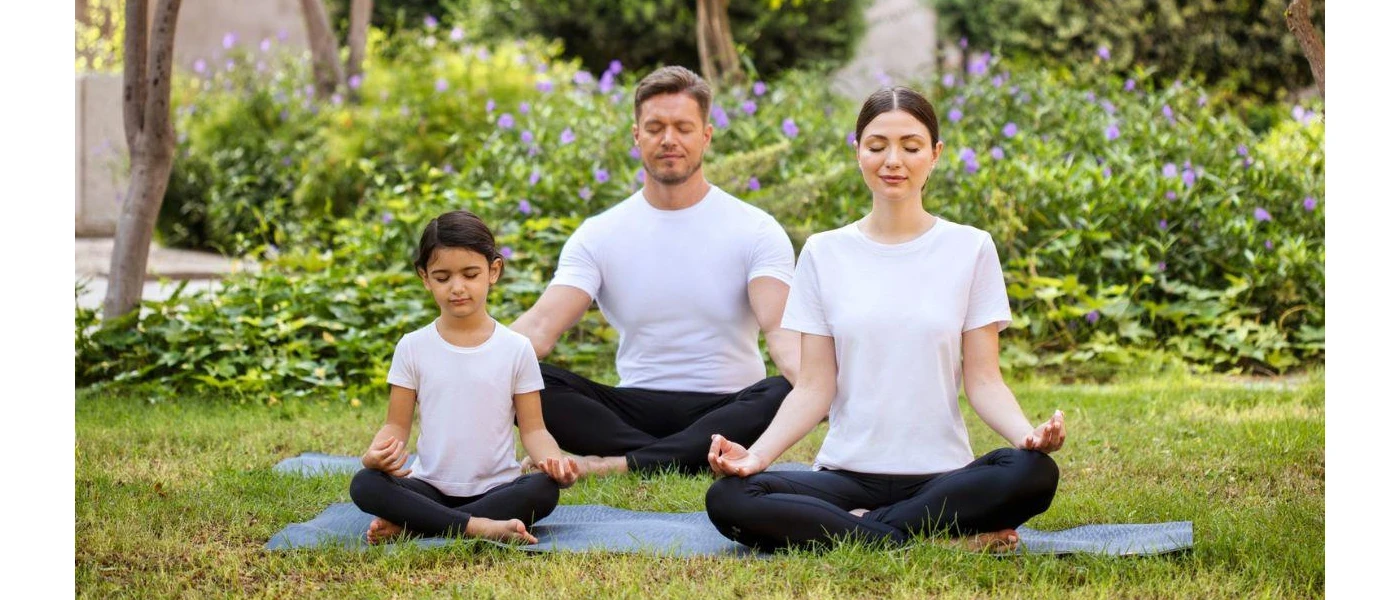 Man, woman and little girl in white t shirts and black trousers meditate on a lawn in a garden