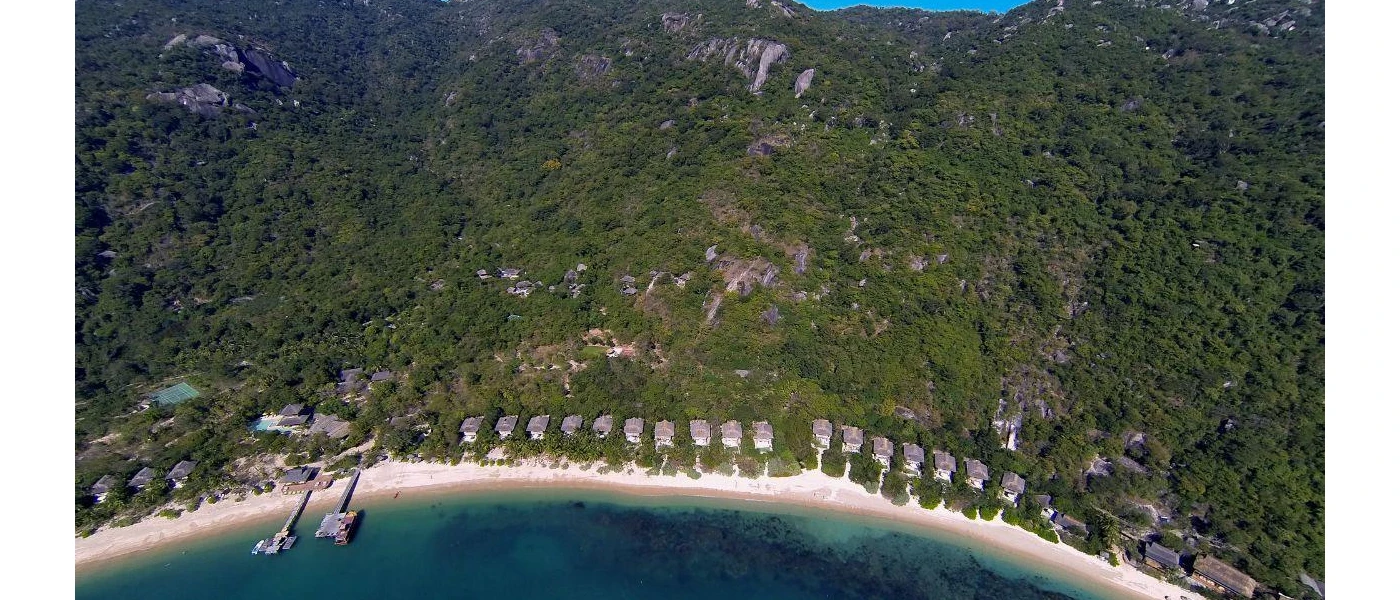 Green-cloaked hillside with a white sandy beach and sea at the bottom, with rows of thatched-roof villas lined up towards the shoreline