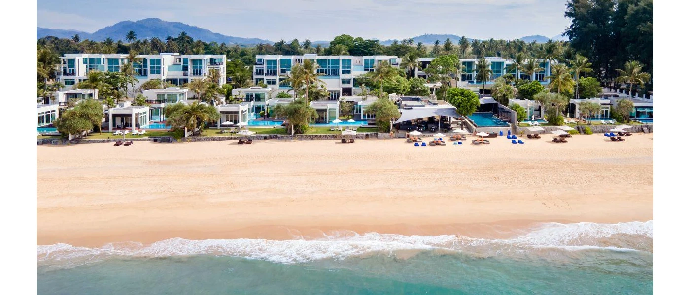 Low-rise white buildings lined up along a beach with private pools and floor-to-ceiling windows, surrounded by palm trees