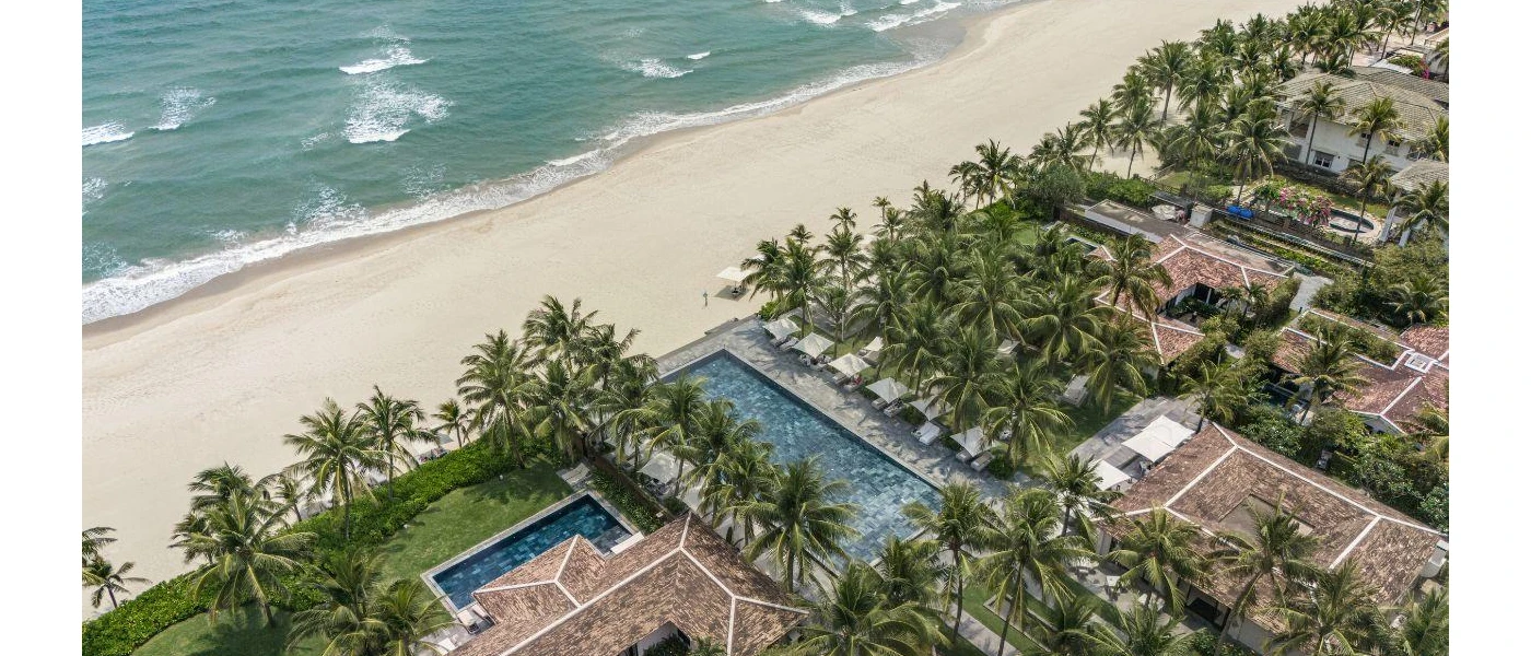 A beachfront resort covered in palm trees overlooking ivory sands, with tile-roofed buildings and swimming pools as seen from above
