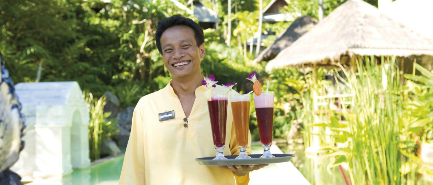 Smiling staff member in yellow carries a tray of tall, colourful drinks through tropical grounds