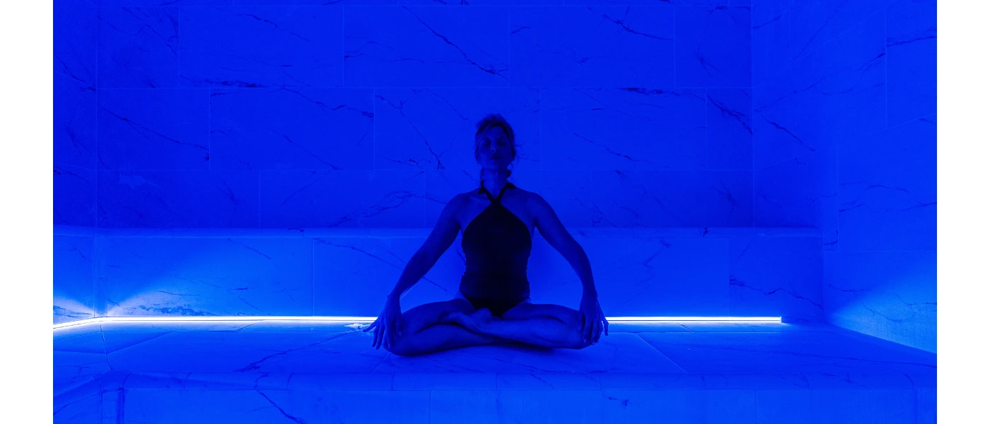 Woman in a black swimsuit sits cross-legged in a blue-lit room