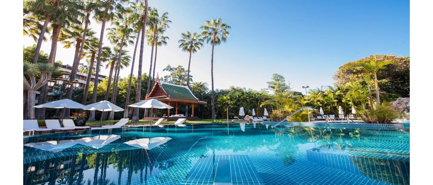 Large turquoise swimming pool surrounded by white parasols and loungers, tall palms and tropical greenery under a blue sky