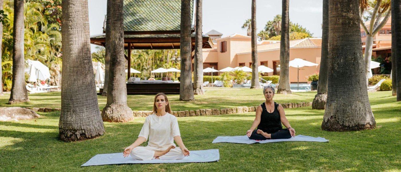 Two women sit in yoga poses on blue mats in a tropical garden
