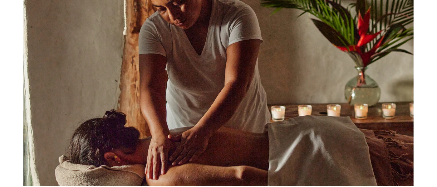 Therapist massages a female lying on her front on a treatment bed in a room with a unit of candles and a vase of flowers