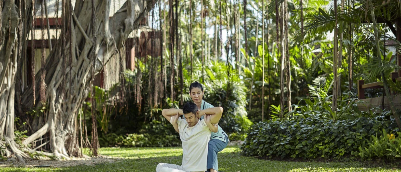Man in white, loose fitting clothes has his arms stretched behind him by an expert in a tropical garden setting