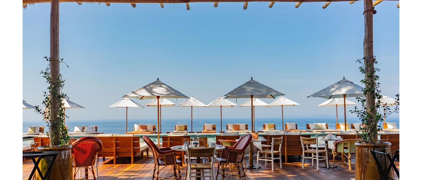 Wooden terrace with tables and chairs, white parasols and a view of the sea in the background